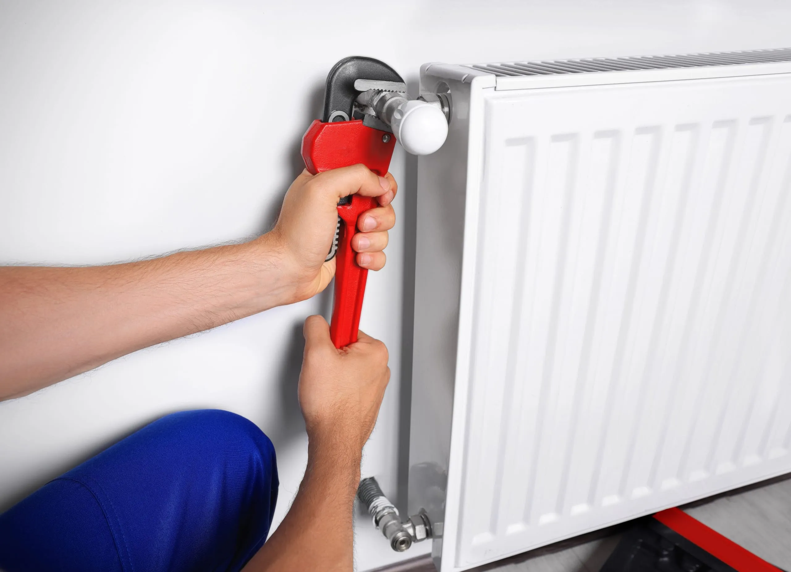 Plombier en tenue bleue serrant à la clé à tuyau rouge le robinet thermostatique d'un radiateur blanc